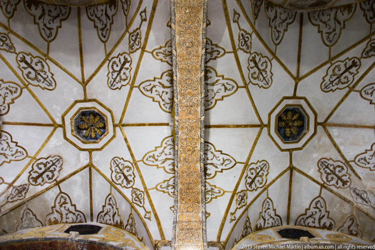 Close view of two pale vault bays with gold ribs, stylized motifs, octagonal bosses, and an ornate dividing band.