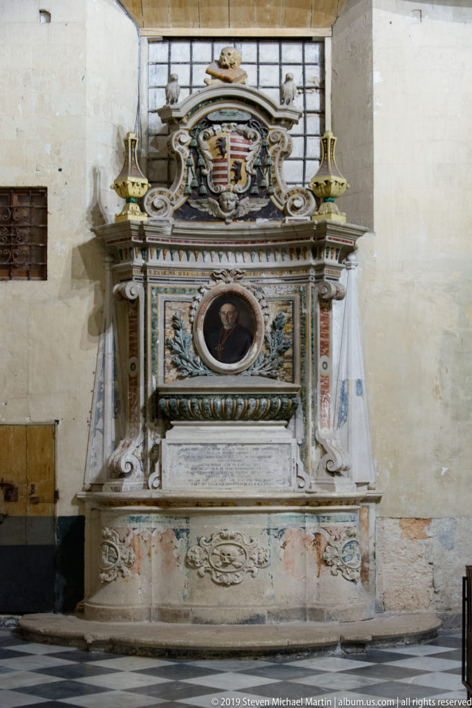 Polychrome wall monument with oval portrait, heraldic crest, inscription tablet, scrollwork, and marble-like architectural frame.
