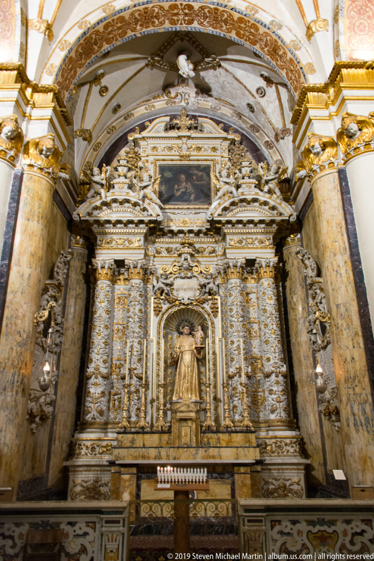 Tall gilded side altar between marble columns, centered on a standing saint in a niche beneath a framed painting.