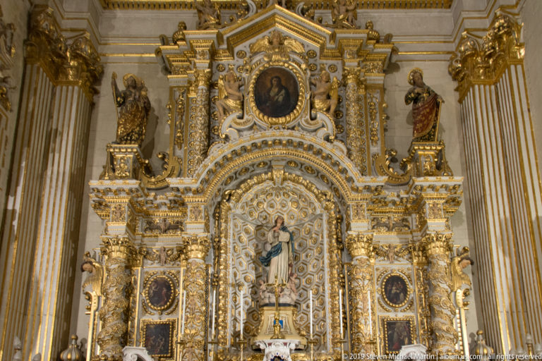 Upper portion of an ornate gilded altar with Marian statue, oval portrait, standing saints, columns, and dense sculptural decoration.