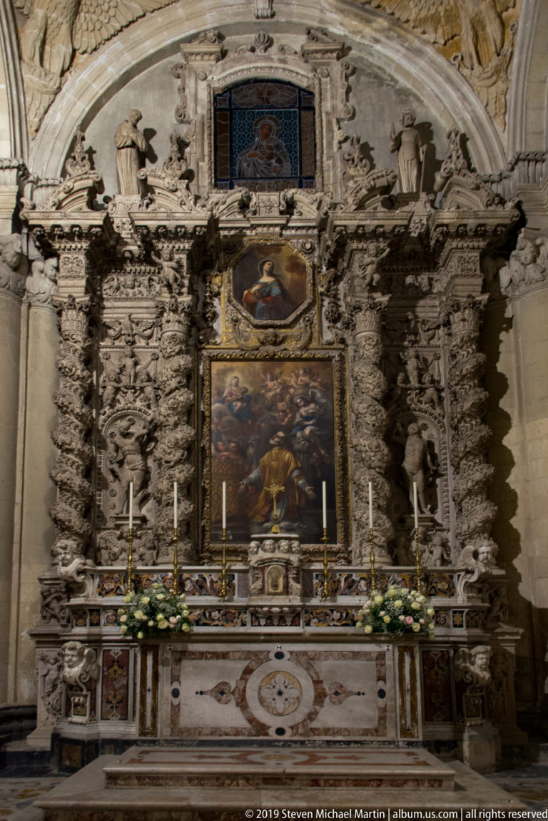 Carved pale-stone side altar with stacked religious paintings, sculptural figures, candles, and flower arrangements.