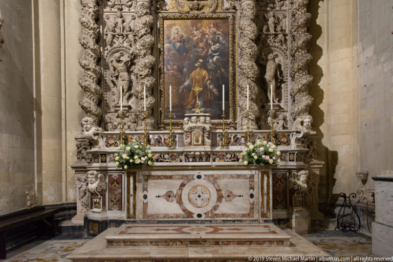 Tall religious painting above a polychrome marble altar, framed by dense pale-stone carving and flanked by flower arrangements.