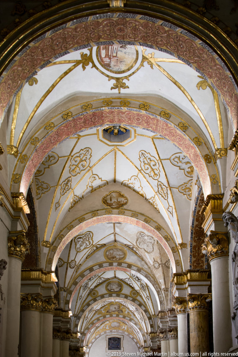 Long perspective beneath repeating white-and-gold vaulted bays with painted motifs, reddish arches, and gilded column capitals.