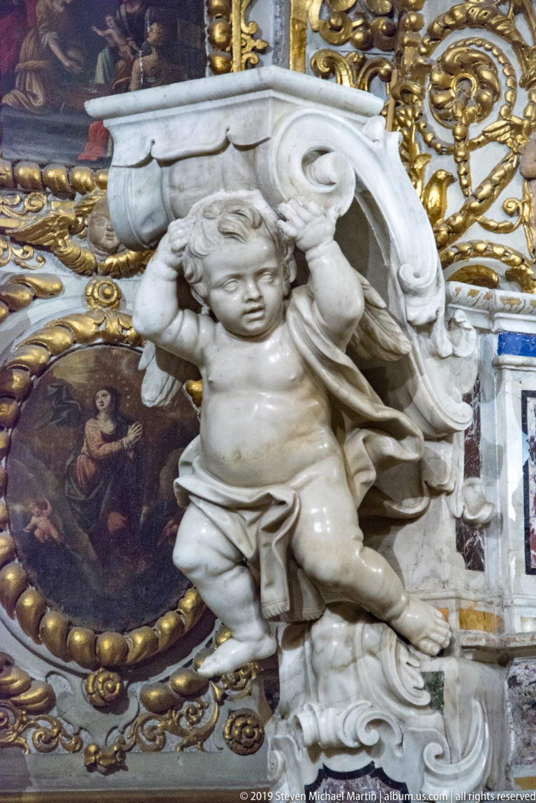 White marble cherub with wings and drapery beneath a curling scroll, set against gilded ornament and an oval painting.