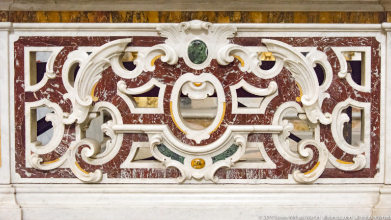 Close view of red marble panel with symmetrical white scrollwork inlay and small green and ochre stone accents.