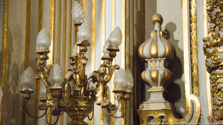 Brass multi-arm light fixture with flame-shaped bulbs beside a striped gold-and-white finial and gilded wall ornament.