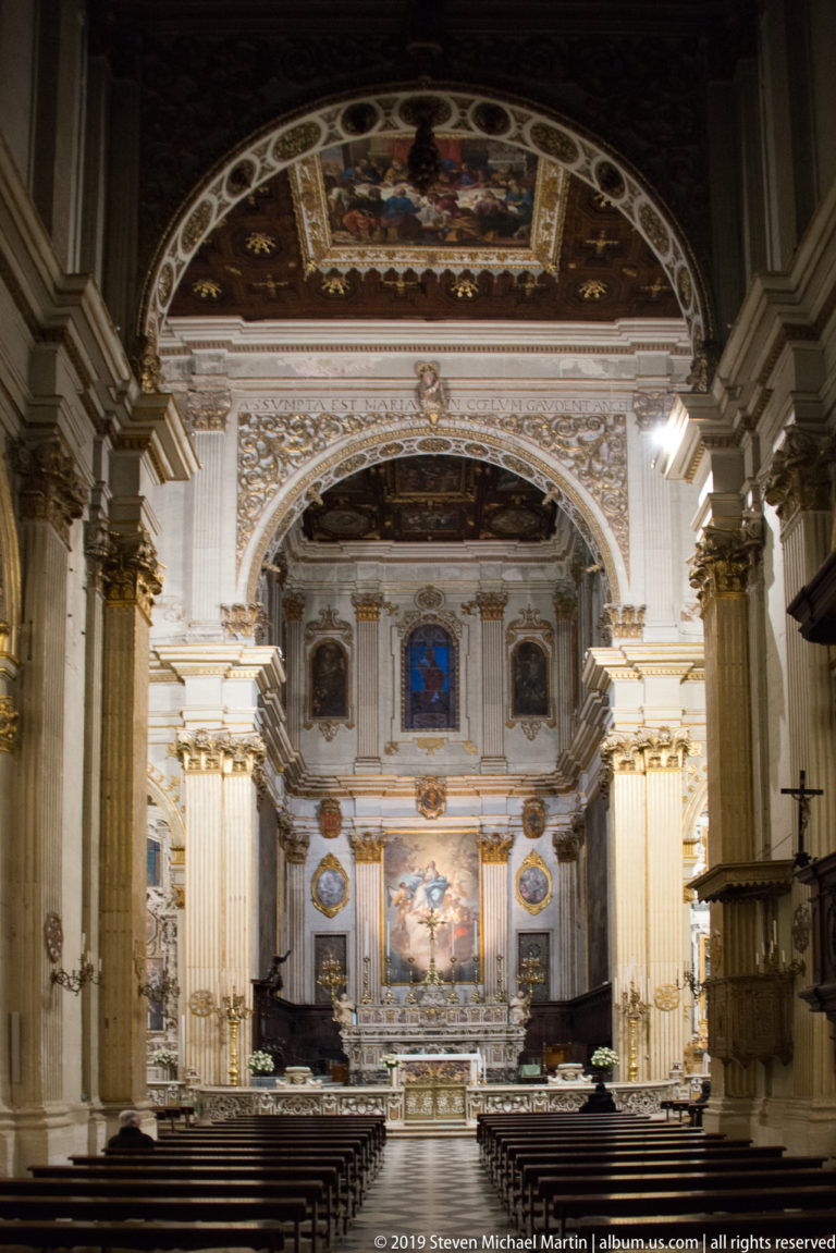 Central nave lined with pews and massive piers, looking through decorated arches toward the brightly lit high altar.