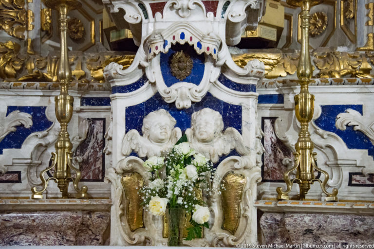 Two white cherubs beneath a blue cartouche on an ornate blue, gold, and marble altar, with candles and white flowers.