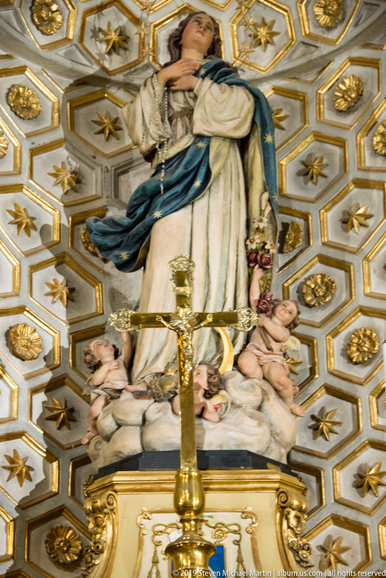 Statue of the Virgin Mary in white and blue above putti, with a brass crucifix and gilded coffered background.