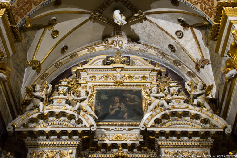 White-and-gold side altar with sculpted figures and a central religious painting beneath a vaulted church ceiling.