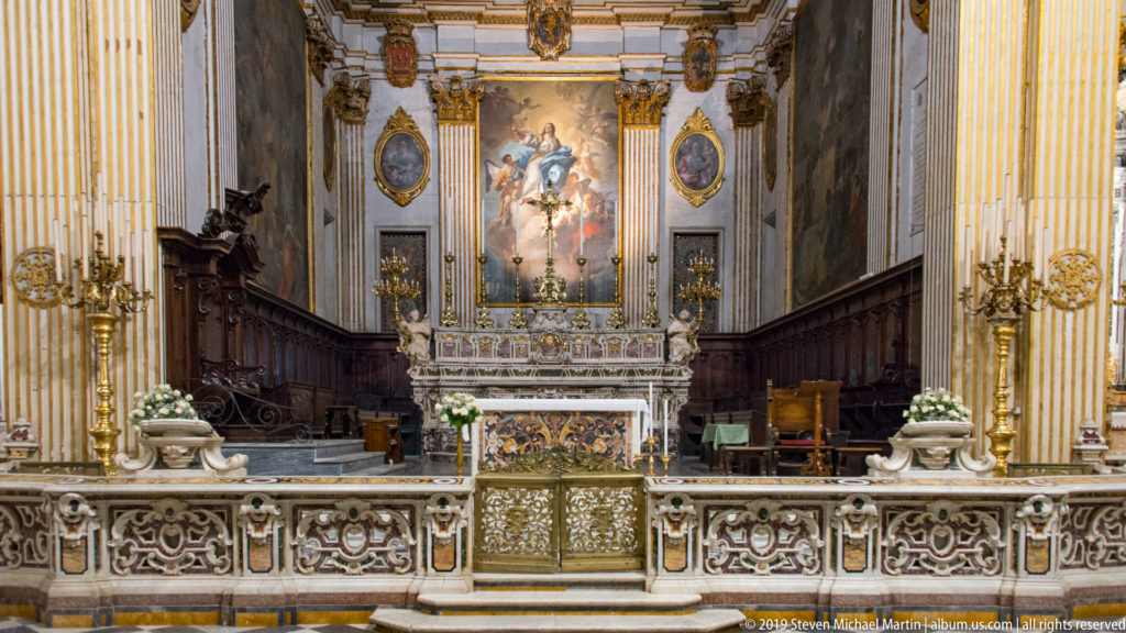 Symmetrical cathedral chancel with ornate altar, dark wooden choir stalls, decorative screen, gilded details, and white flowers.