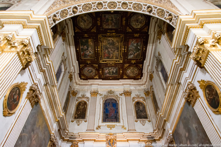 Upward interior view of a dark painted coffered ceiling above white-and-gold walls, framed paintings, and stained glass.