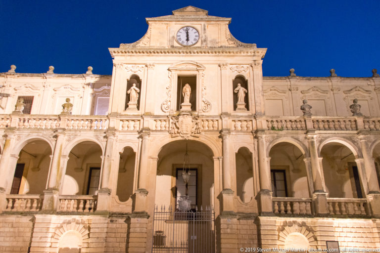 Illuminated stone façade with central clock, statues in niches, arcades, balustrades, steps, and a gated entrance at night.