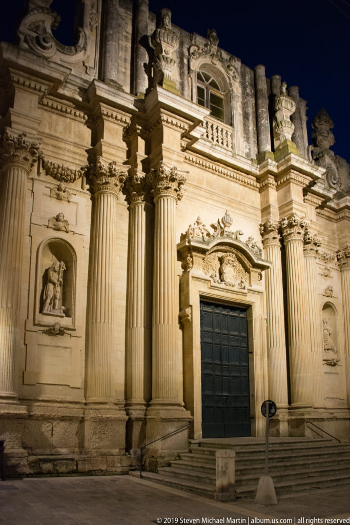 Oblique night view of an ornate stone church façade with tall columns, dark doorway, sculptural niches, and layered moldings.