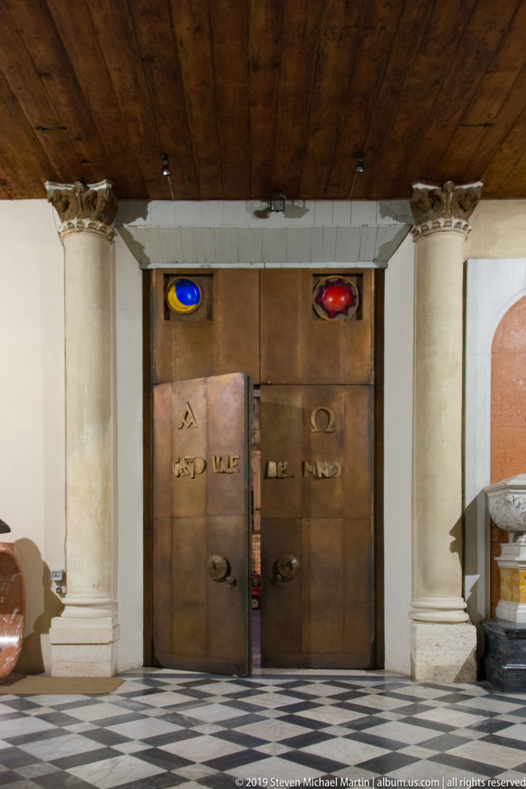 Bronze double doors between stone columns, with colored circular details above and a black-and-white patterned floor in front.