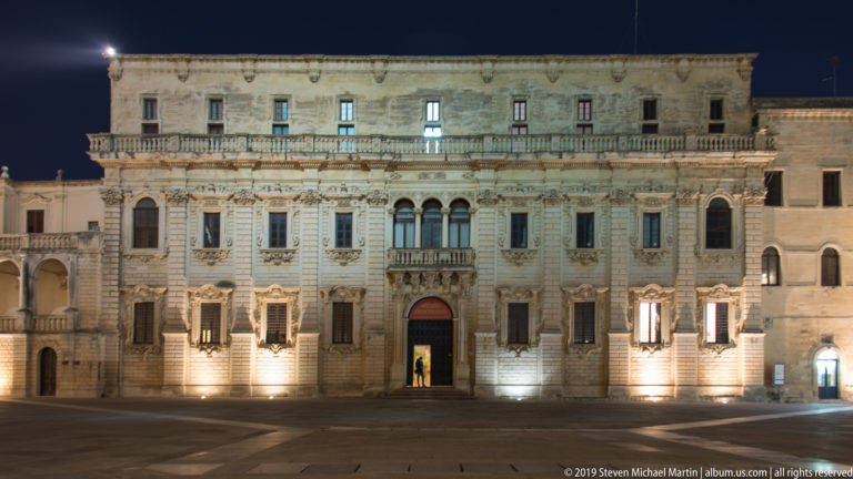 Large illuminated stone palace with rows of windows, balconies, carved ornament, and a central arched doorway facing the square.