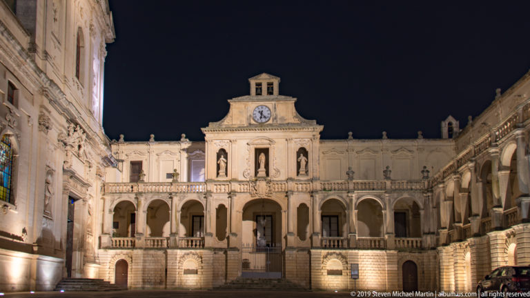 Illuminated pale-stone courtyard façade with arcades, statues, balustrade, central entrance, and clock pavilion at night.