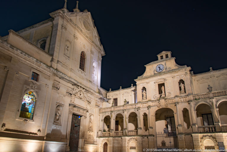 Wide night view of pale stone cathedral buildings around Piazza del Duomo, including stained glass, arcades, statues, and a clock.