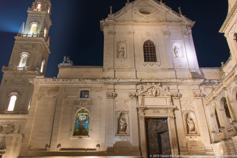 Illuminated cathedral side façade with triangular pediment, central doorway, statues in niches, stained glass, and bell tower edge.