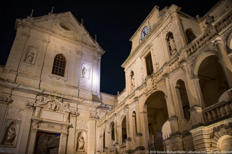 Low-angle night view of cathedral façade with statues and portal beside a clocked arcaded building receding to the right.