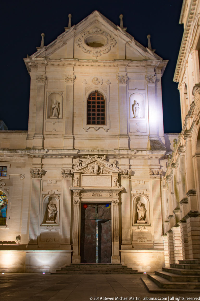 Frontal night view of a pale stone cathedral façade with triangular pediment, central window, dark doorway, and four sculptural niches.