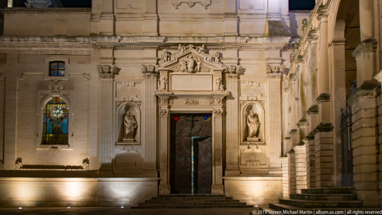 Ornate cathedral doorway with carved stone surround, flanking statues, adjacent stained-glass window, and arcade edge at right.