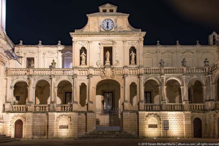 Symmetrical illuminated façade with clock, three statues, central stairs and gate, and long arcades extending on both sides.