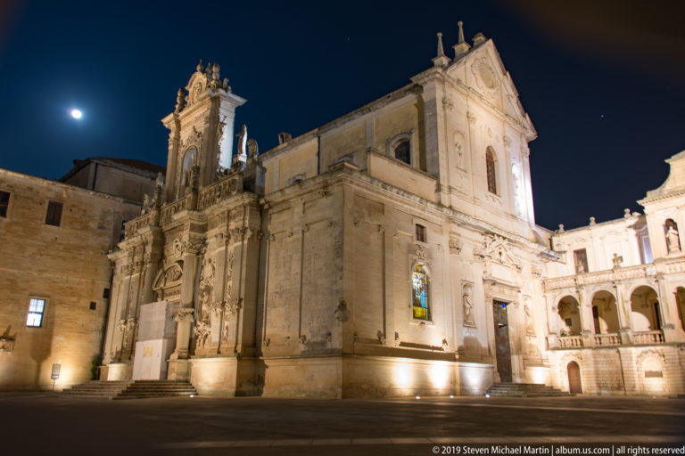 Oblique night view of illuminated cathedral with ornate entrance, long side wall, stained glass, arcades, and moon above the square.