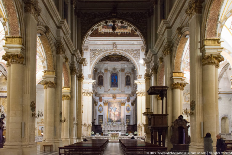 Nave view toward the sanctuary, framed by pale stone piers and arches, with a dark wooden pulpit on the right.
