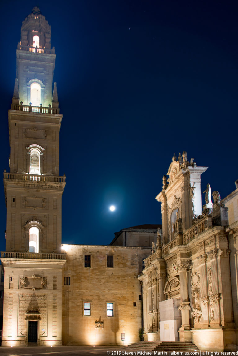 Illuminated square bell tower at left and ornate carved façade at right beneath a bright moon in a deep blue night sky.