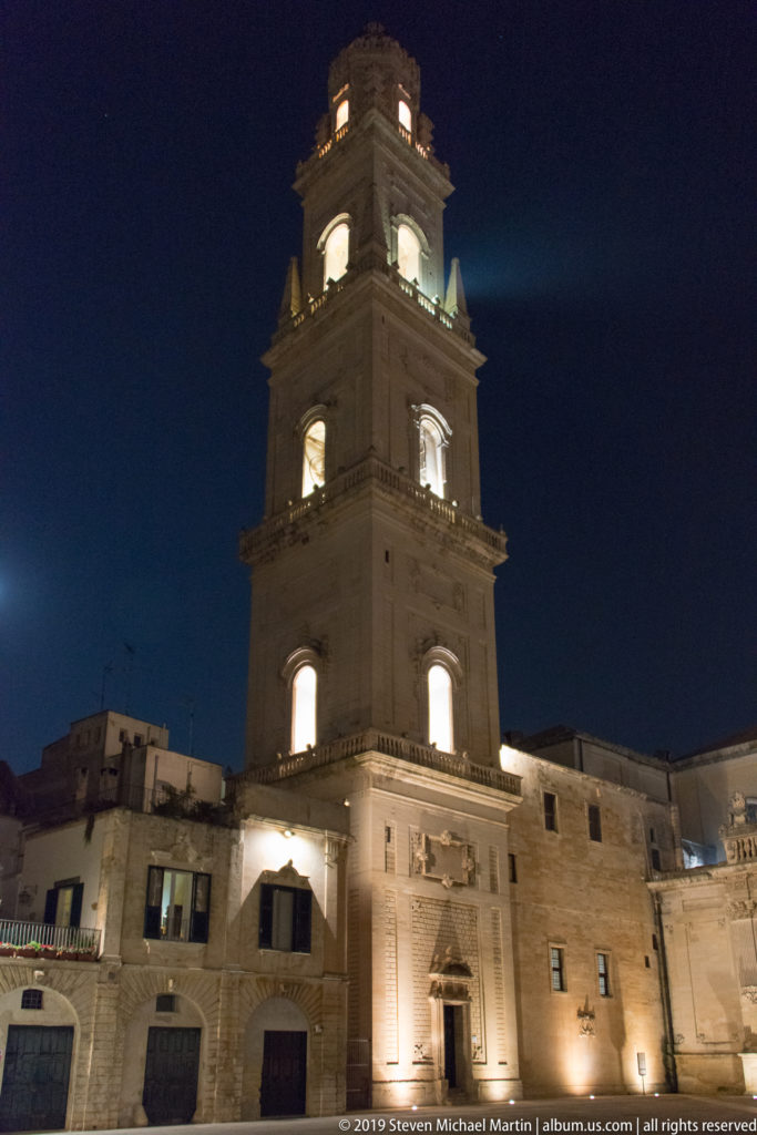 Tall illuminated stone bell tower with stacked square stages and bright openings rising above lower buildings at night.