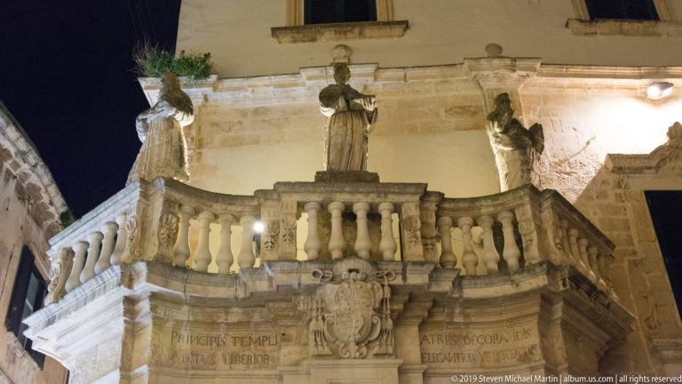 Three stone statues above a curved balustrade and carved coat of arms, with Latin inscriptions on the illuminated masonry below.