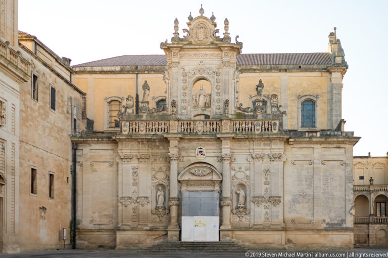 Daylight frontal view of an ornate pale limestone church façade with portal, niches, balustrade, relief carving, and elaborate crest.