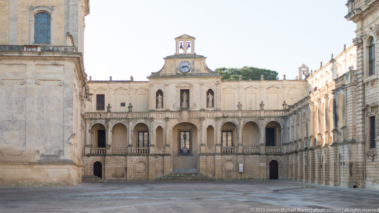 Daylight view across an open stone square toward a symmetrical arcaded façade with central clock, stairs, and balustrades.