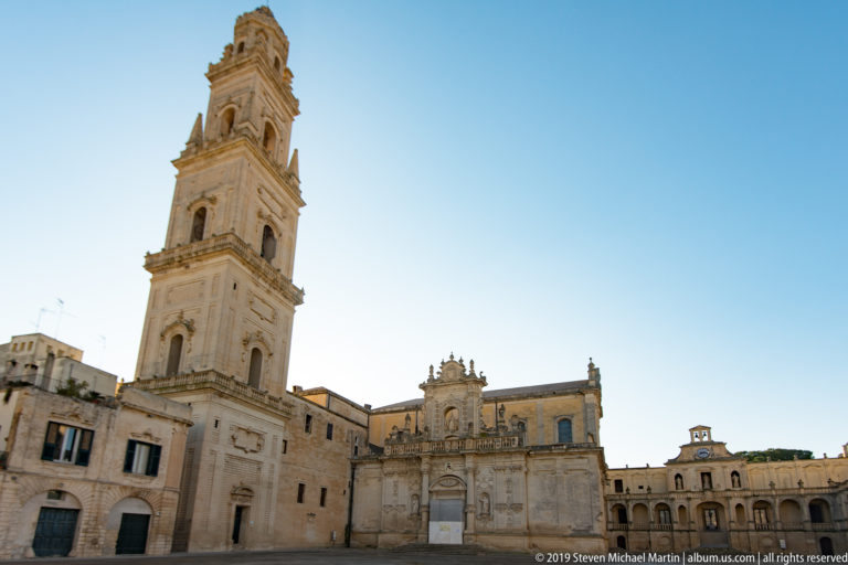 Wide daylight view of tall cathedral bell tower, ornate façade, and arcaded buildings around Piazza del Duomo under blue sky.