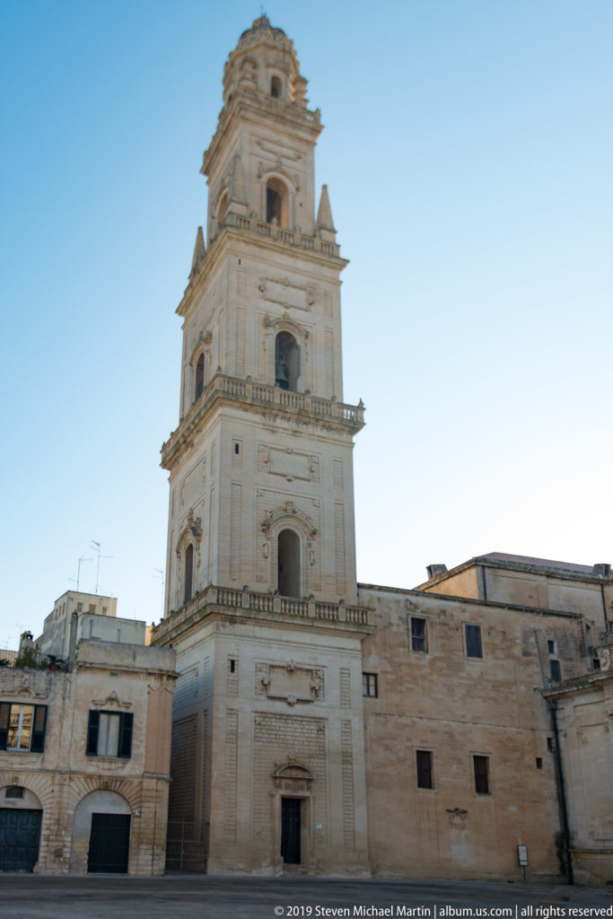 Close daylight view of pale stone bell tower with stacked stages, arched openings, cornices, and balustrades above adjoining buildings.