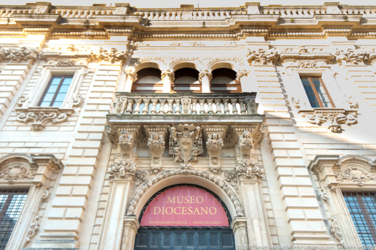 Ornate pale-stone Museo Diocesano entrance with an arched doorway, balcony, sculpted figures, balustrade, and pink museum sign.