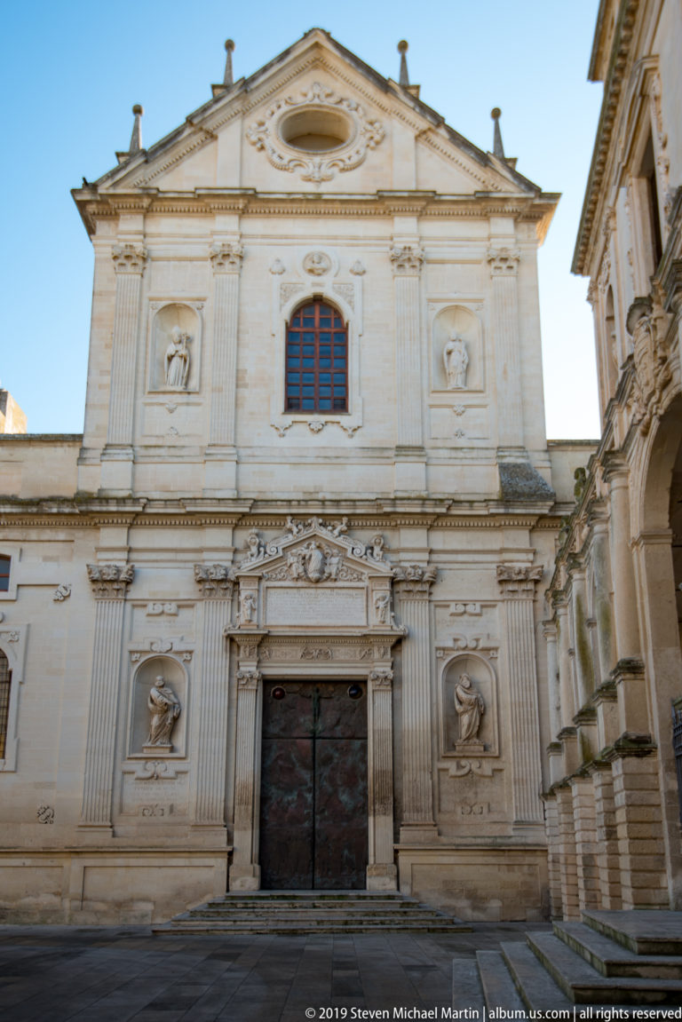 Frontal daylight view of pale cathedral façade with triangular pediment, central window, carved doorway, pilasters, and four niches.