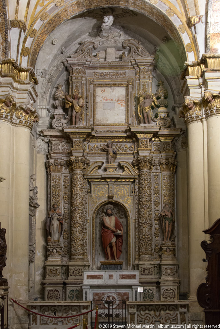 Highly ornamented stone side altar with seated saint in a central niche, carved figures, columns, and inscribed panel above.