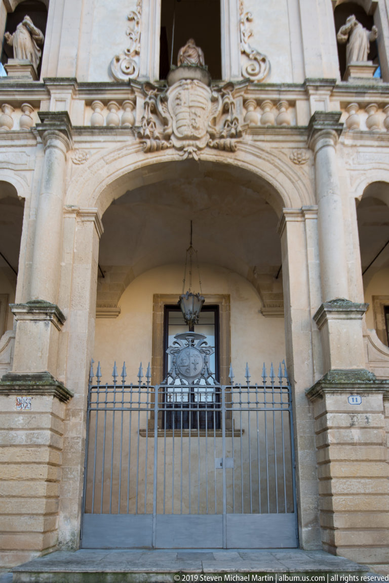 Wrought-metal gate beneath a broad carved stone arch with hanging lantern, heavy piers, and heraldic ornament above.