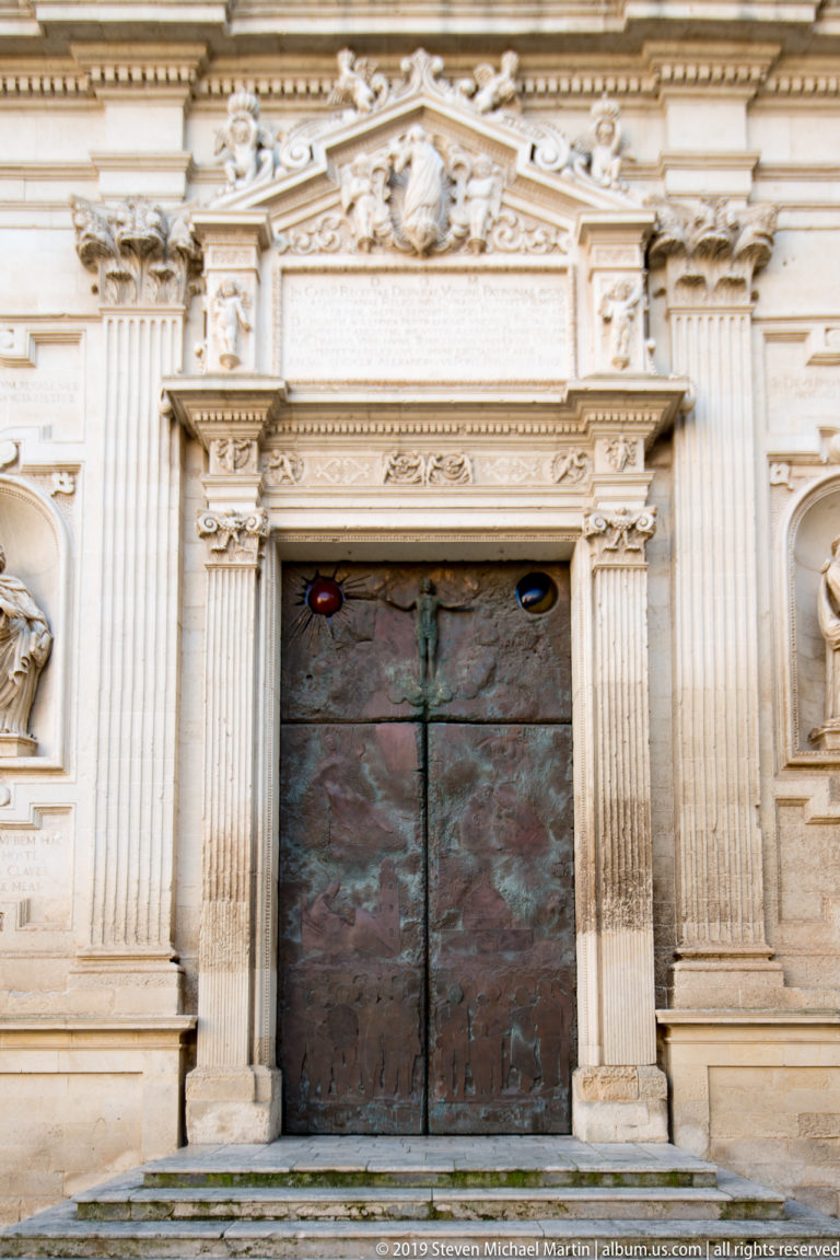 Dark double doors within an ornate pale stone portal with fluted pilasters, carved reliefs, inscription panel, and small colored lights.