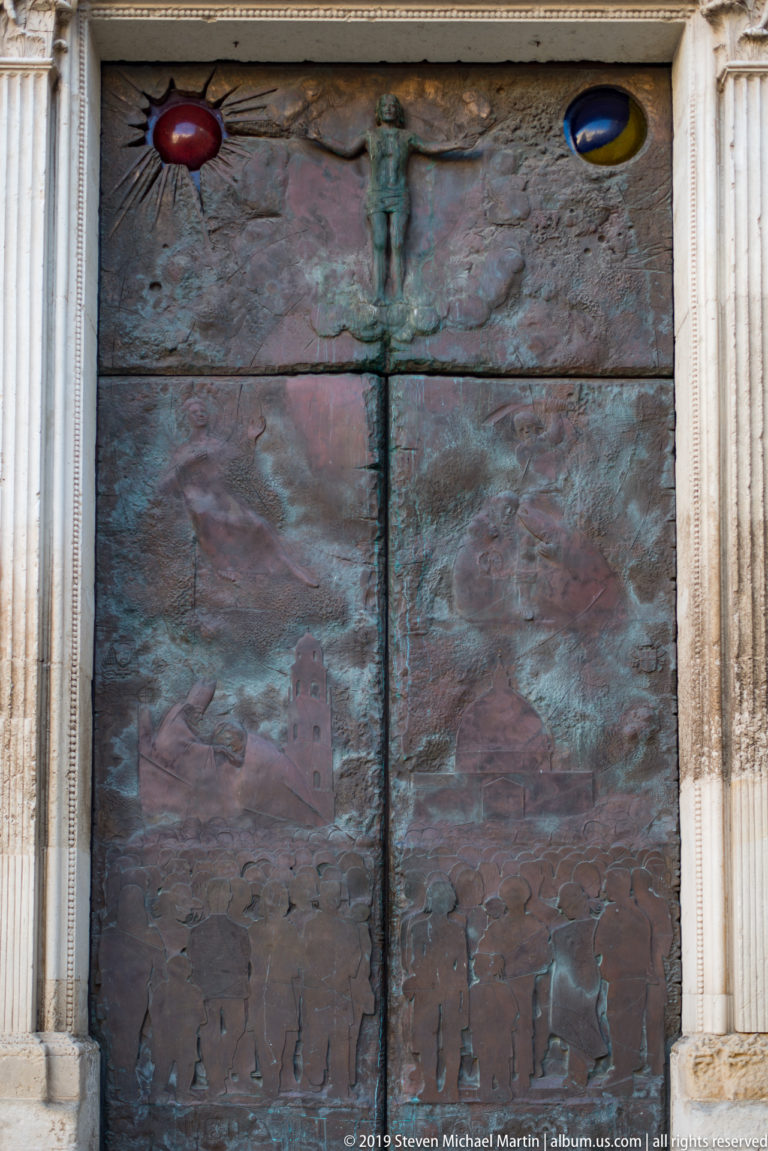 Bronze church door with a crucified figure, textured relief panels, colored circular insets, and pale carved stone surround.