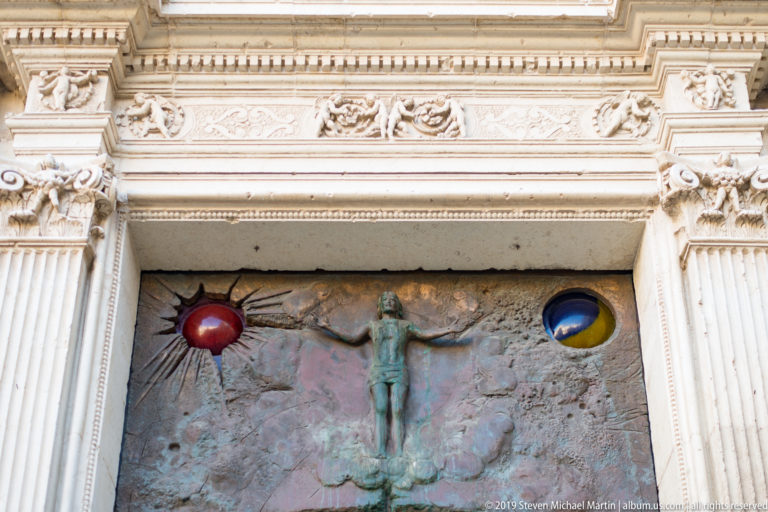 Upper bronze door panel with a central crucified figure, red and blue-yellow circular insets, and an ornate pale stone frame.