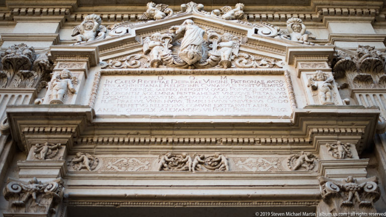Latin inscription panel framed by carved stone moldings, relief ornament, projecting cornices, and a sculptural group above.
