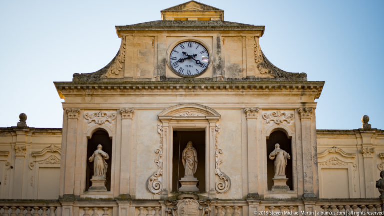 Upper façade of Lecce’s Archbishop’s Palace with a central clock, the Virgin and two flanking statues in niches, and carved Baroque stonework.