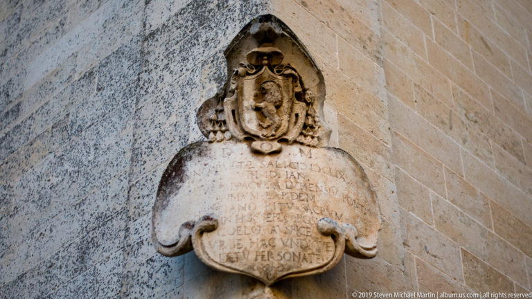 Weathered stone memorial plaque with a heraldic crest and worn Latin inscription mounted on a masonry wall.