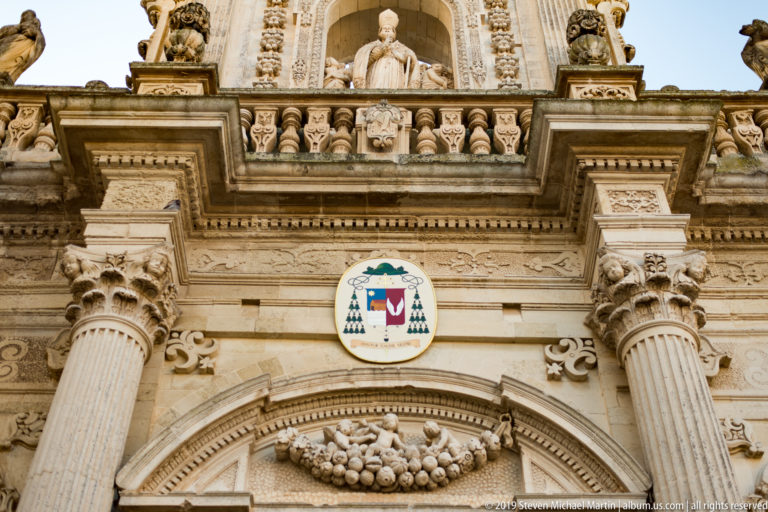 Ornate stone portal with fluted columns, episcopal coat of arms, fruit garland, balustrade, and seated clerical statue above.