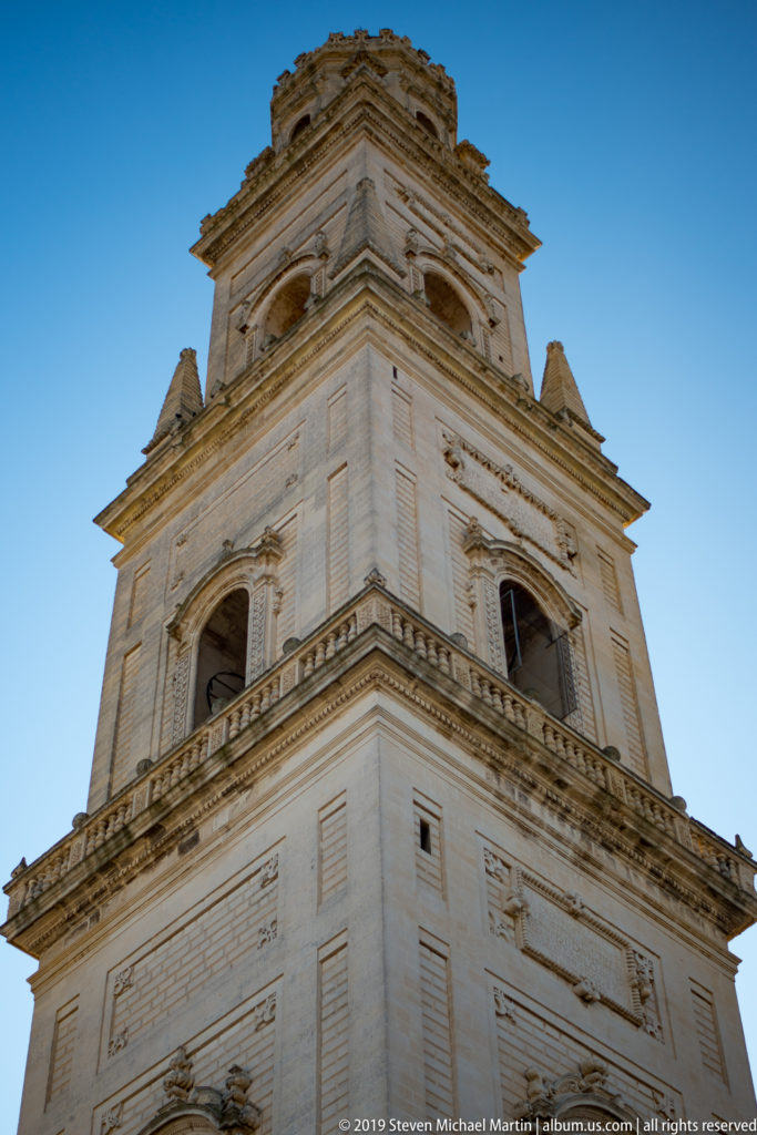 Upward view of Lecce Cathedral bell tower with stacked pale-stone stages, projecting cornices, pilasters, and arched bell openings.