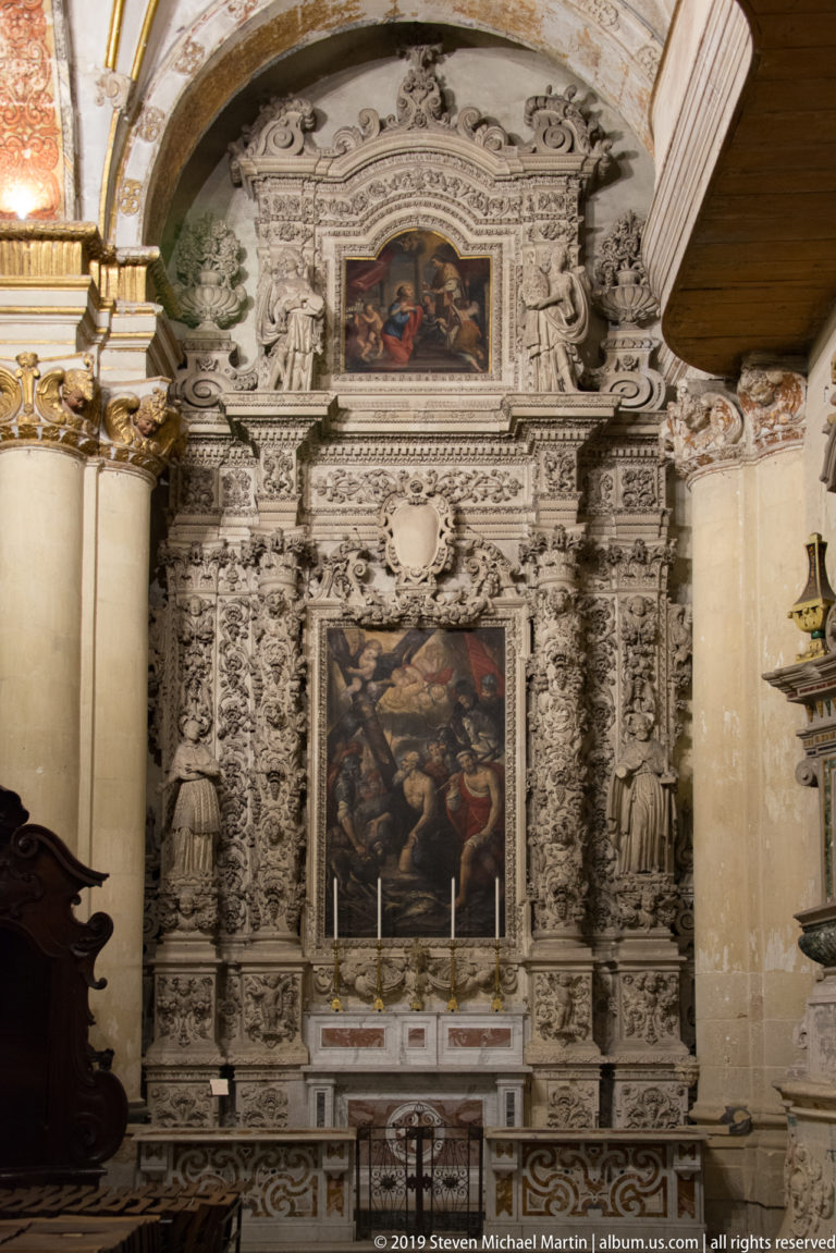 Ornate pale-stone side altar with dense sculptural carving and two vertically stacked religious paintings.