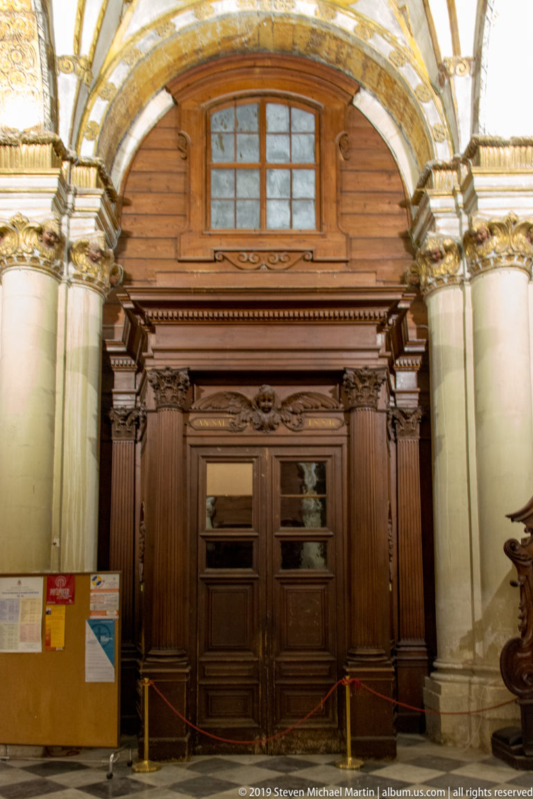 Tall dark wooden double doorway with paneled surround and cornice, set between stone columns beneath a glazed opening.